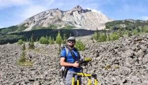 A research stands in front of a huge rock glacier with trees around it.