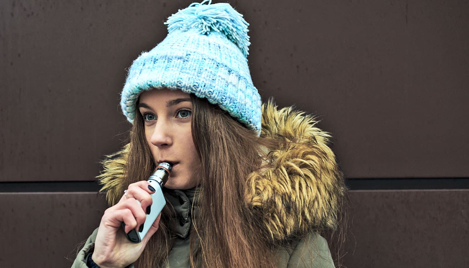 A young woman in winter clothing vapes outside.