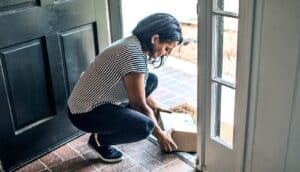 A woman kneels down to pick up a package from her front doorstep.