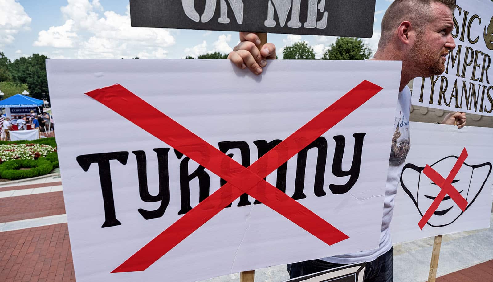 man holds mask and "tyranny" signs with red tape Xs