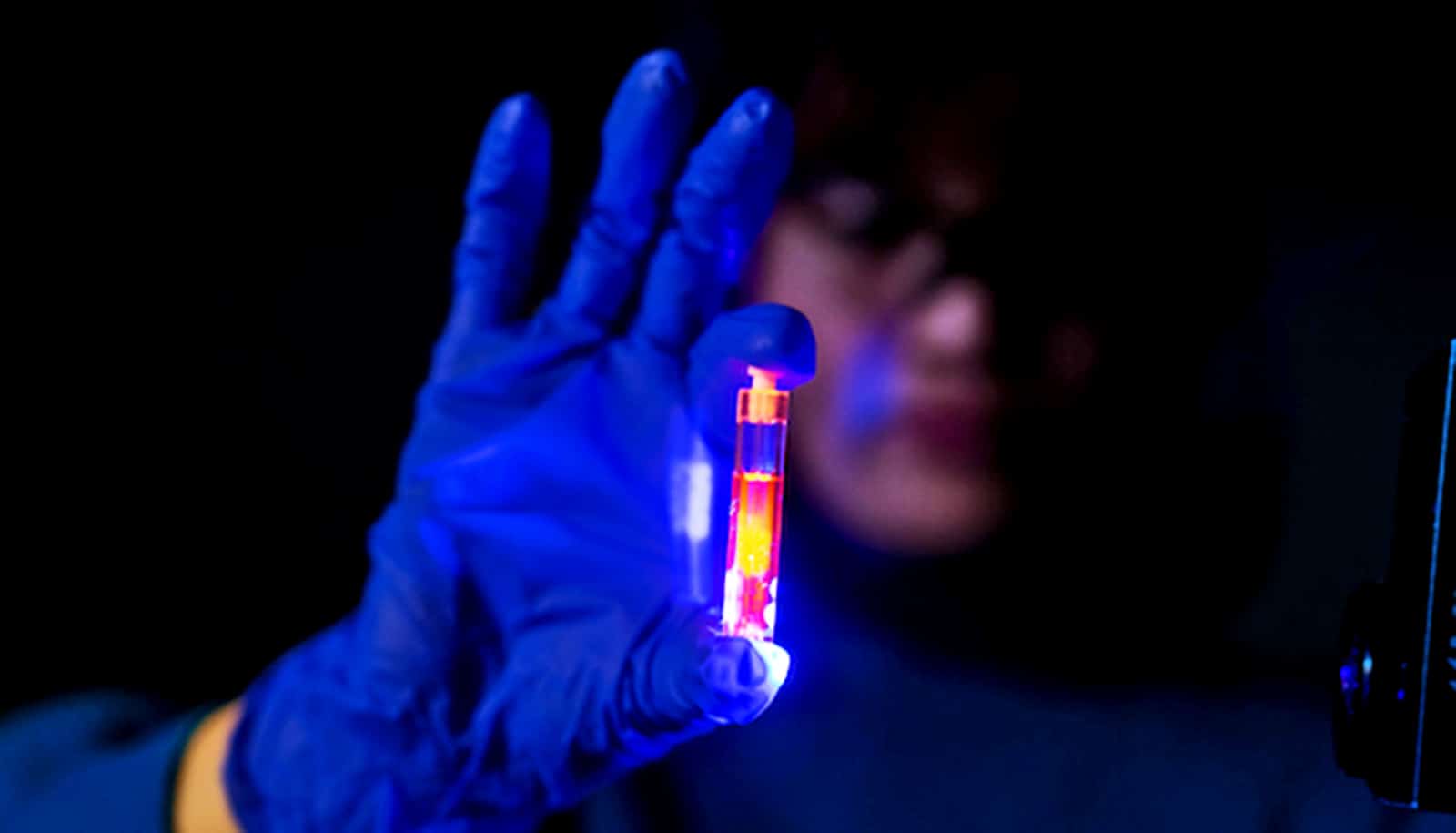 A researcher wearing blue gloves holds up a glowing orange vial in a dark room.