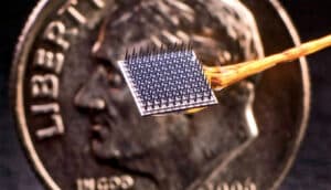 The BrainGate implant is a tiny metallic square with what look like many pins sticking out of its surface. A penny is shown much larger than the implant in the background for scale comparison.
