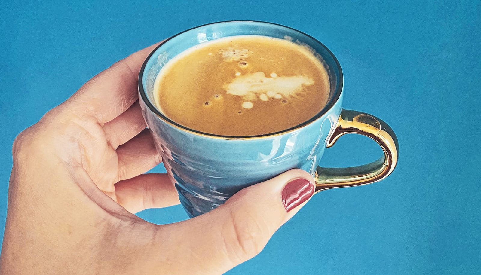 Eine Frau hält eine blaue Tasse Kaffee mit goldenem Henkel vor blauem Hintergrund.