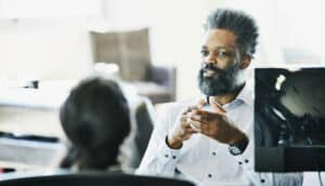 Black man in dress shirt speaks to person at desk with customer monitor