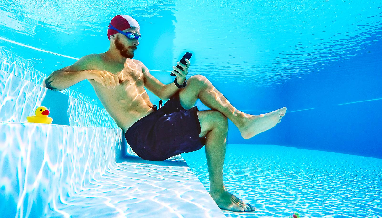 A man in a swimming cap and goggles sits underwater in a pool while looking at his phone.