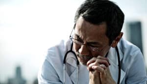 A doctor looking down while resting his face on his clasped hands.