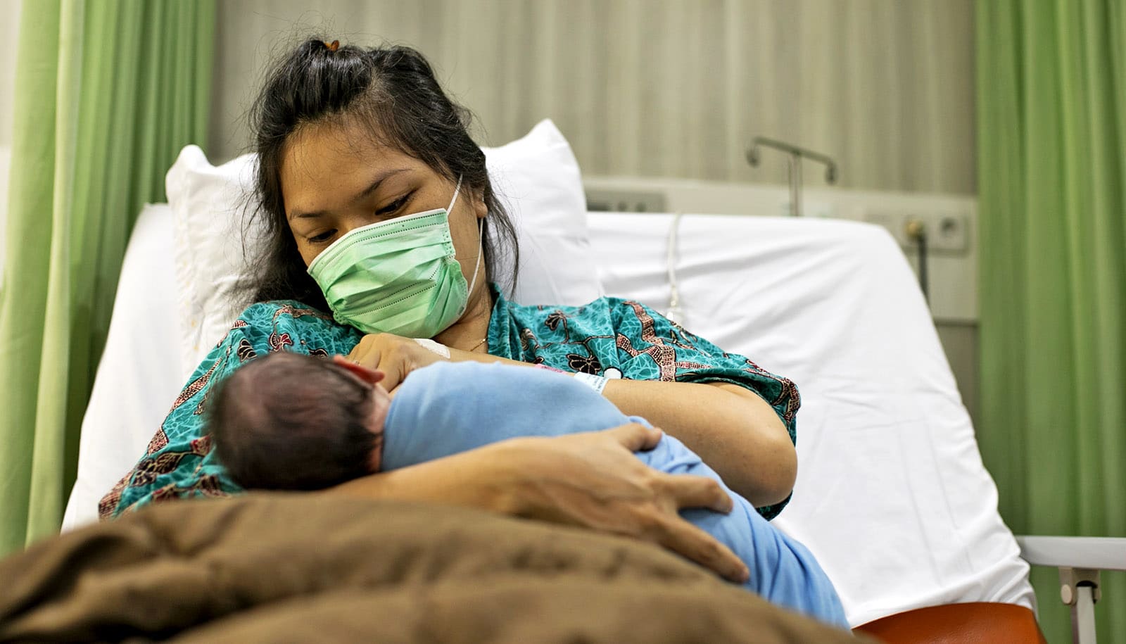 adult in mask holds newborn baby in hospital bed