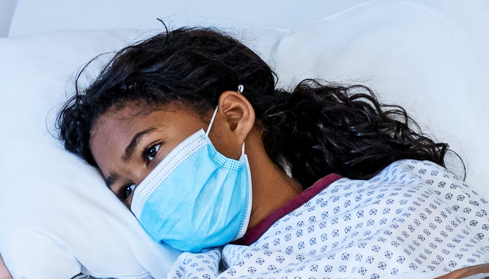 A young girl lays in a hospital bed wearing a blue face mask.
