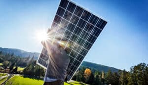 A man carries a solar panel up a hill with a forested mountain range in the background.