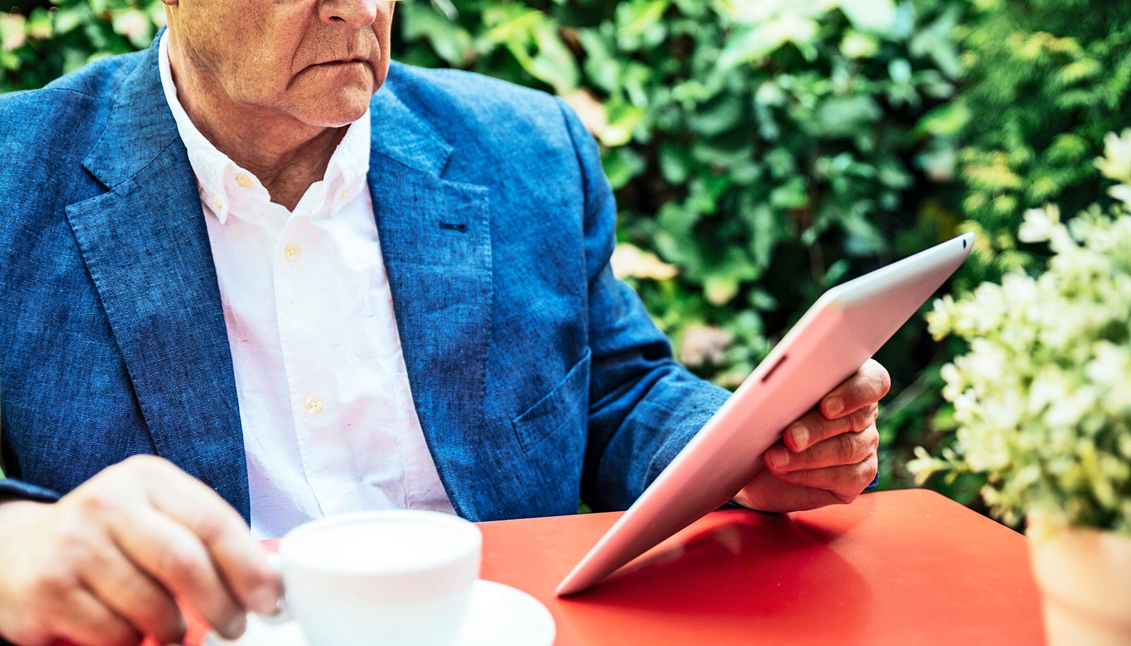 A man in a white shirt and blue blazer reads a tablet computer while drinking coffee outdoors.