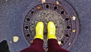 A person wearing bright yellow sneakers stands on a sewer hole cover on the street.