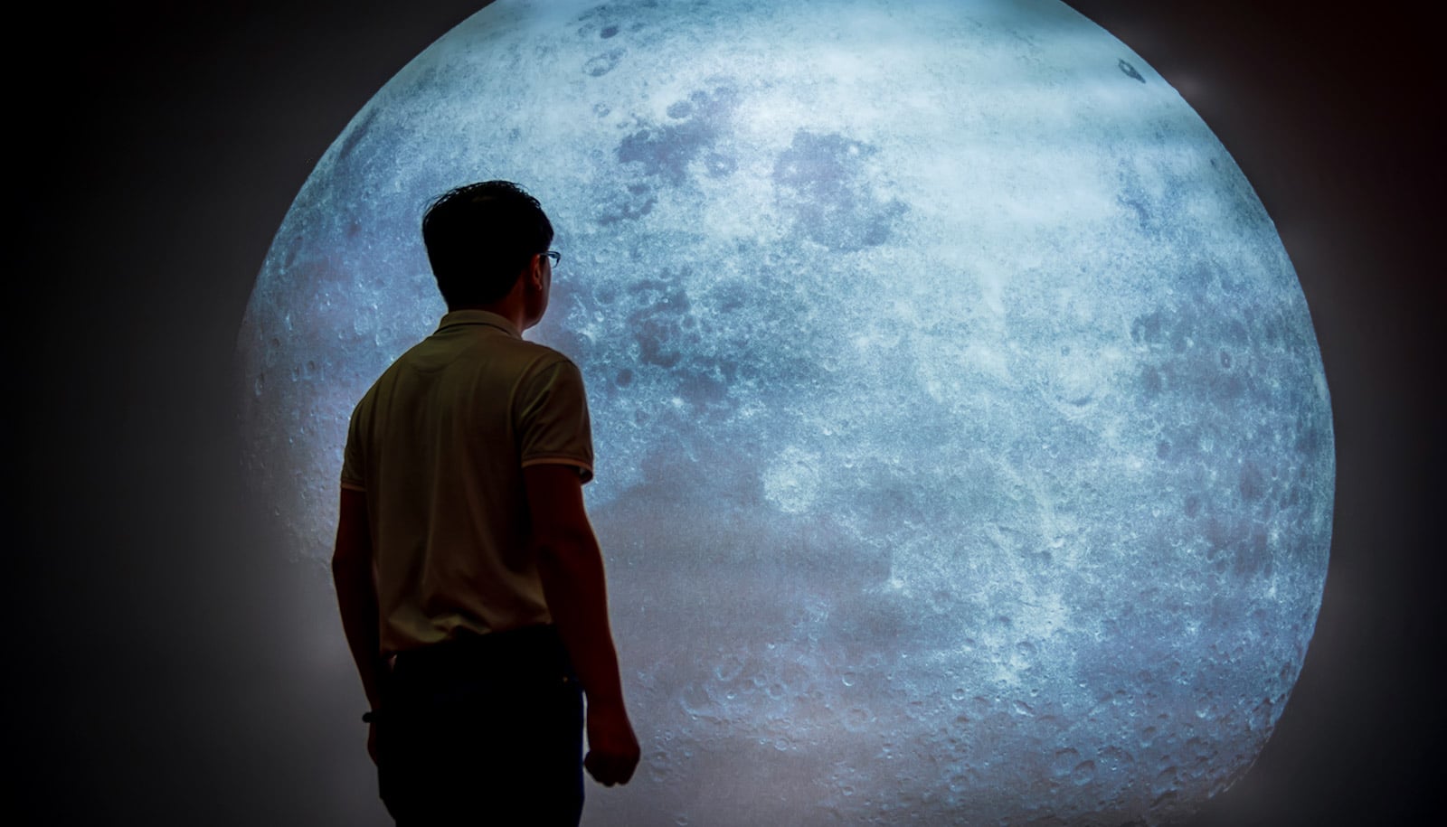 A man looks at a projection of the moon on the wall of a dark room.