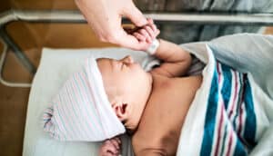 A mother holds her baby's hand while the baby lays in a hospital bassinet.
