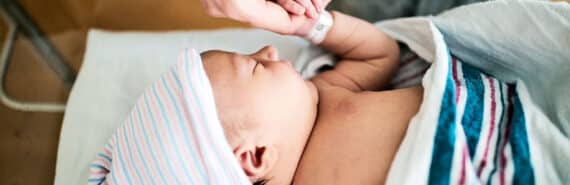 A mother holds her baby's hand while the baby lays in a hospital bassinet.