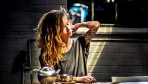 A woman reaches up to her neck while sitting at a kitchen table.