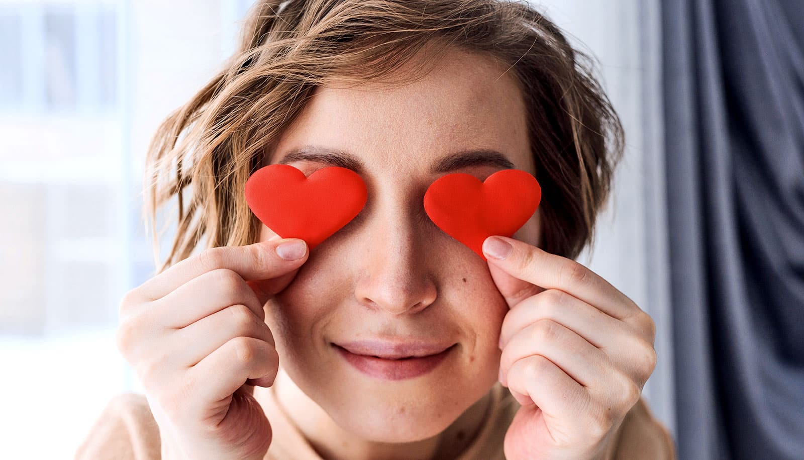 A woman holds heart-shaped pieces of paper over her eyes.