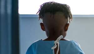 A young boy in a hospital gown sits on the edge of a bed.
