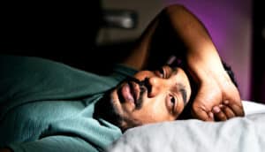 A young man in bed has his arm over his forehead and looks very tired.