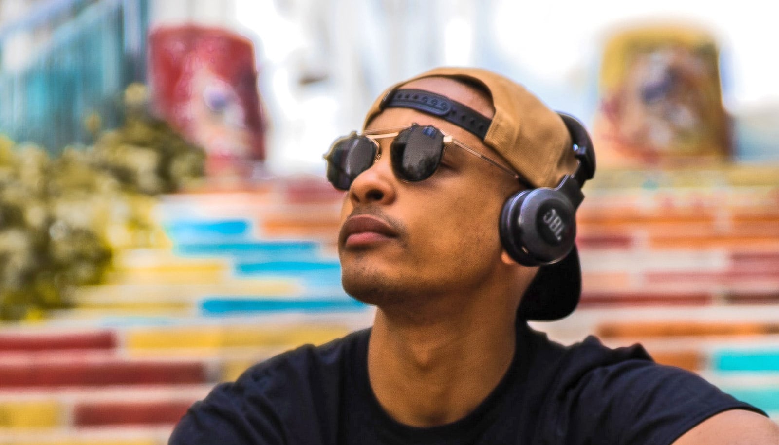 A young man wearing sunglasses, a ball cap, and headphones sits on colorful stairs outside.