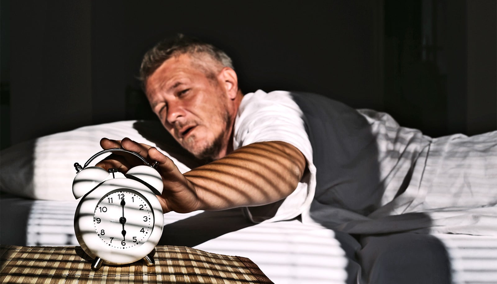 A man reaches from his bed to his alarm clock while the room is still dark.