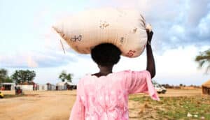 A woman carries a bag of food on her head down a dirt road.