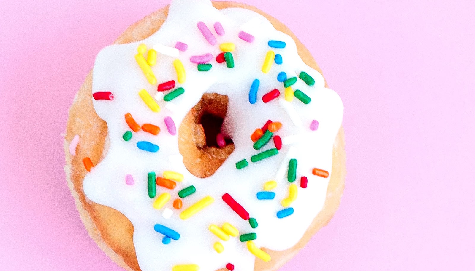 A frosted donut with sprinkles on it on a pink background.