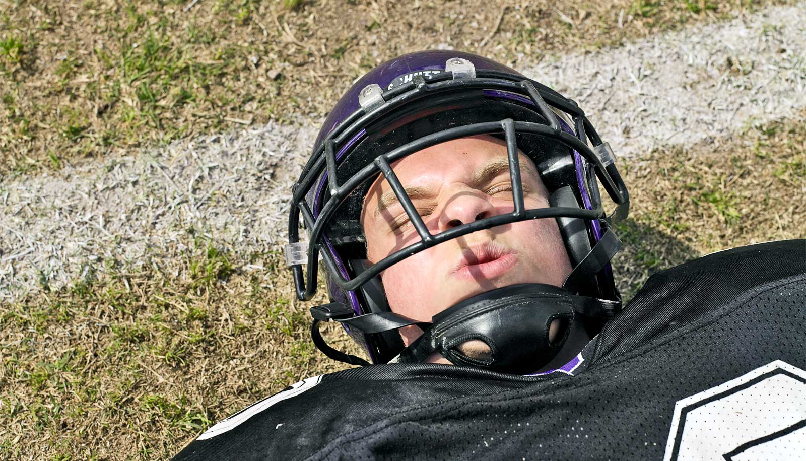 En ung fodboldspiller iført hjelm og uniform grimasserer i smerte, mens han ligger på banen.