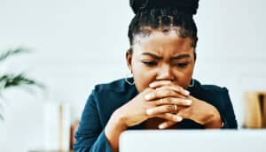 A woman furrows her brown and leans on her clasped hands while reading from a laptop.