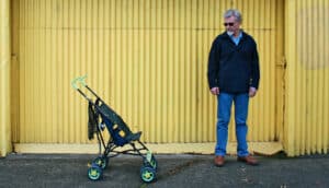 older adult stands awkwardly in front of yellow wall with baby stroller