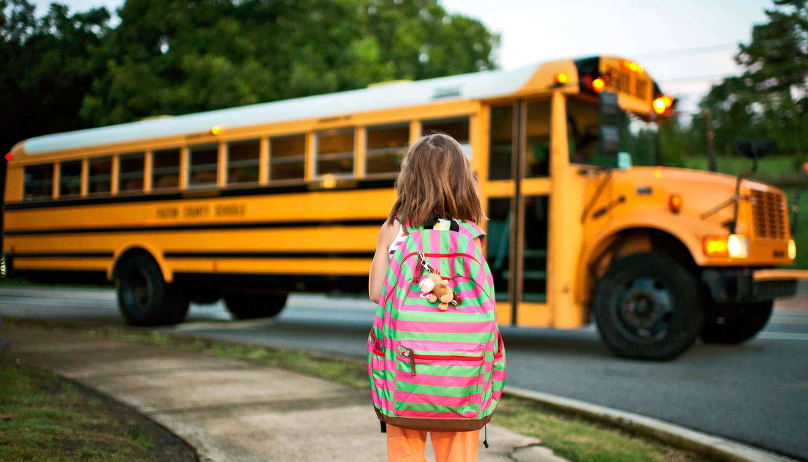 A young girl stands waiting on the sidewalk as a yellow school bus pulls up.