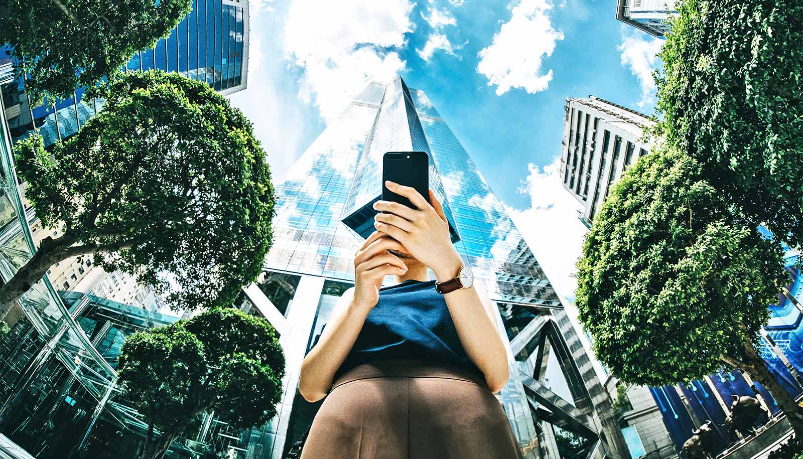 A woman looks down at her phone as trees and buildings tower over her in the background.
