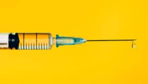 A syringe with liquid coming out of the needle against a yellow background.