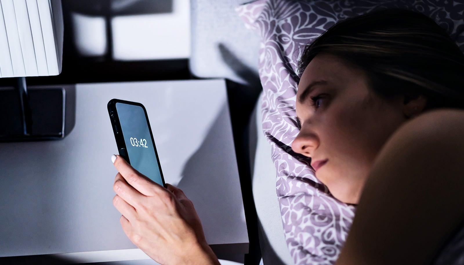 A woman laying in bed looks at the time on the phone on her bedside table.