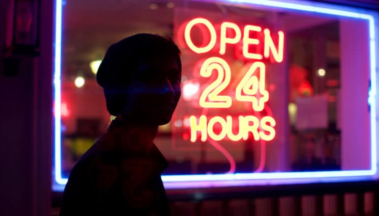 upper body of person in silhouette in front of neon sign that says "open 24 hours"