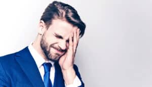 A man in a blue suit and tie grimaces and touches his face with his hand.