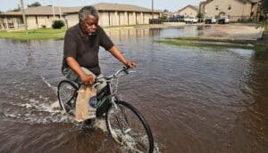 A man rides his bike through flooded streets while carrying a plastic bag of groceries.