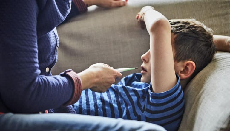 adult holds thermometer in front of child lying on couch with arm over eyes