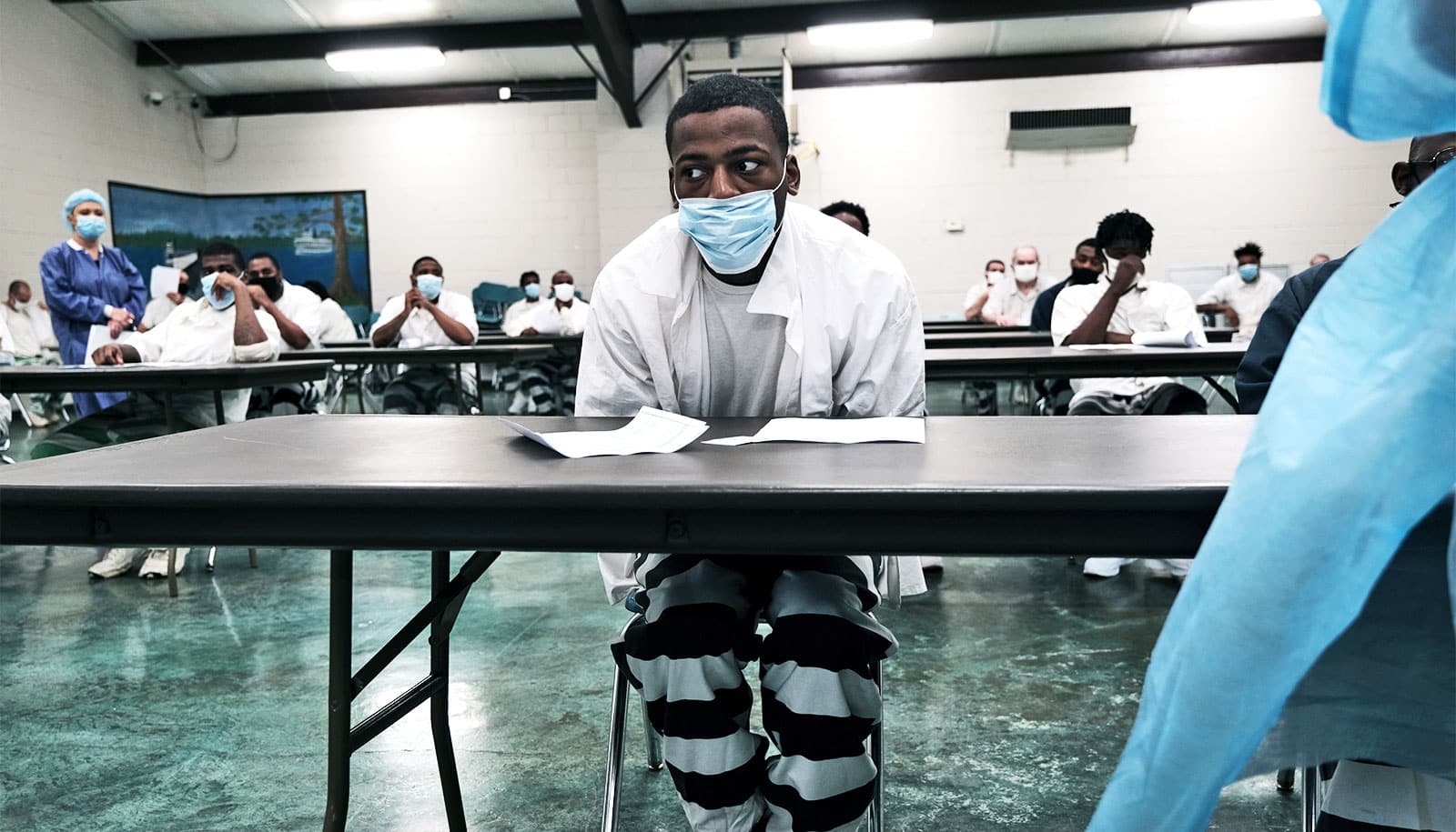 A man sits at a folding table wearing a face mask as he waits with other incarcerated people for a COVID vaccine.