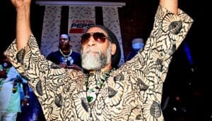 DJ Kool Herc lifts his arms in the air in front of the stage as Busta Rhymes stands behind him holding a microphone.