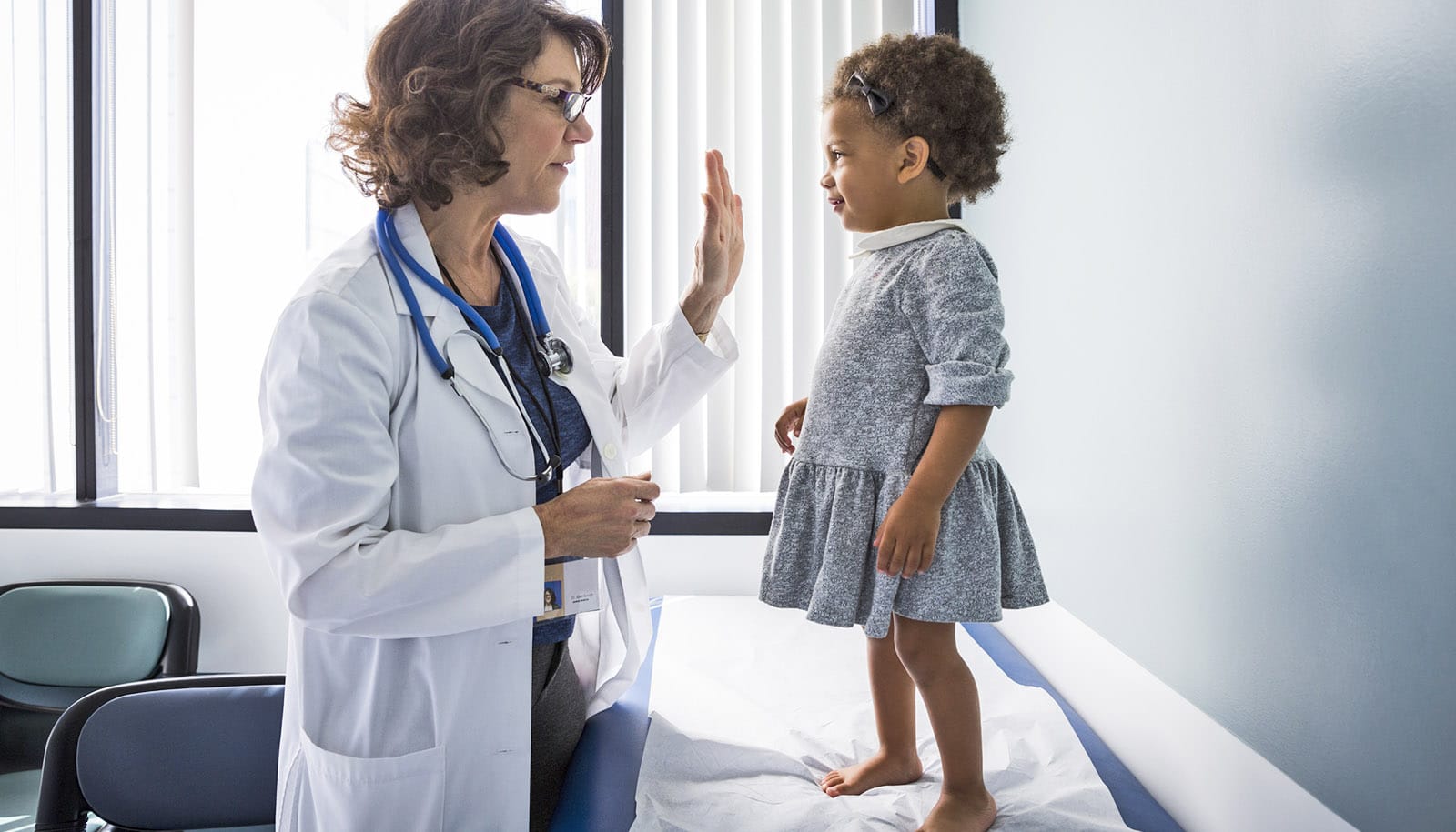 pediatrician holds up hand for high-five to toddler standing on exam table