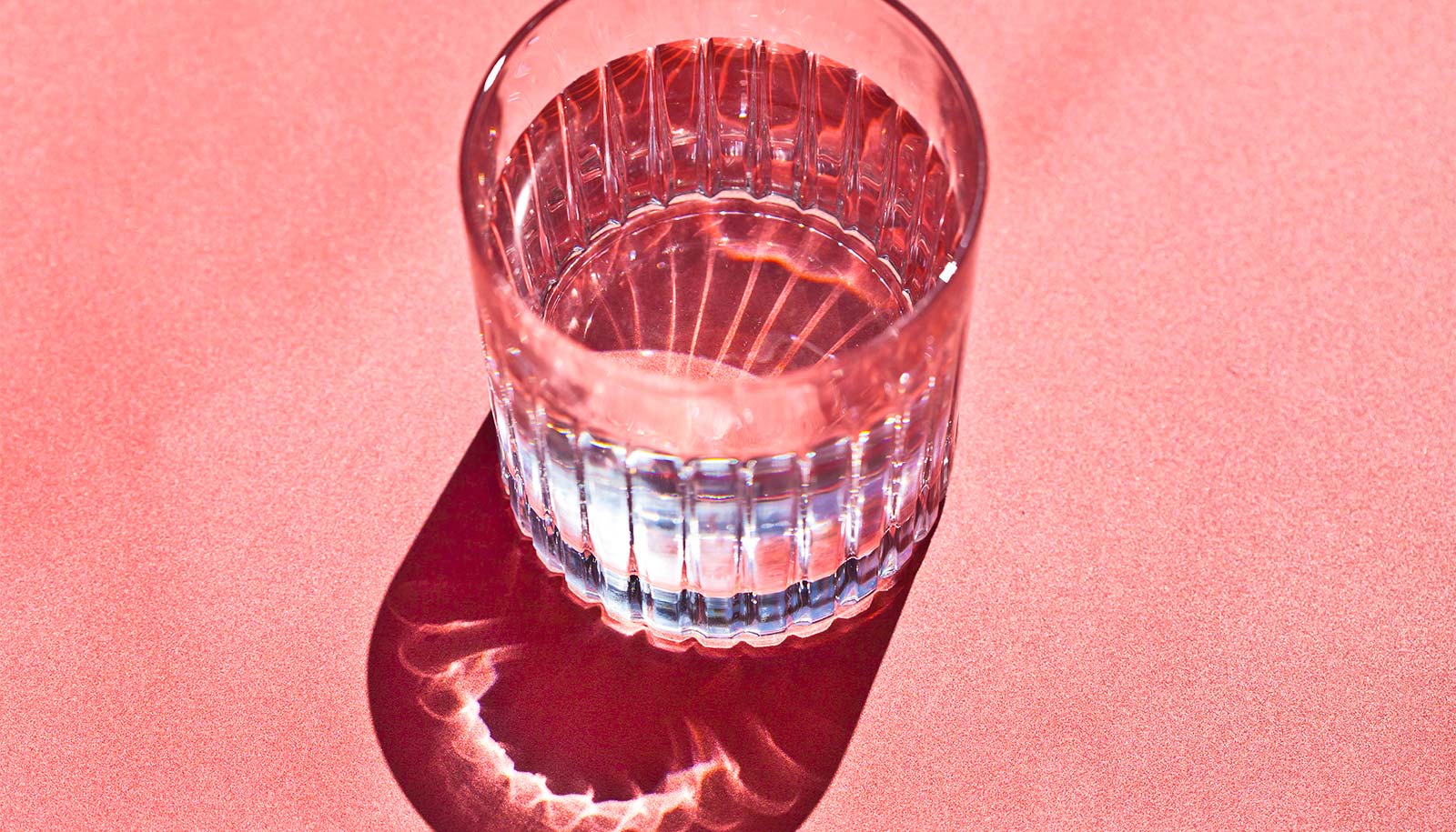 A glass of water with light shining through it on a pink surface.