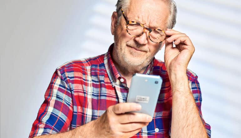 A man in a red, white, and blue plaid shirt looks down with frustration at his phone.