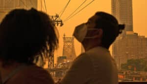 A man wears a mask in the foreground with New York City behind him shrouded in orange smoke.