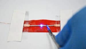 A thin piece of the stretchable material sits on a white surface held down with tape while a research works on it with a small tool.