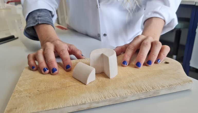 hands rest on cutting board next to small white wheel of "cheese"