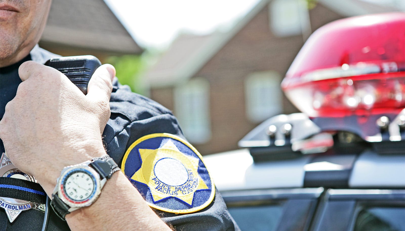 A police officer speaks into his radio while standing in front of a police car and houses.