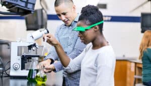 A young woman wearing safety goggles in a science lab works with a pipette while her teacher looks on.
