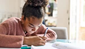 A student studies at home at a table near a window.