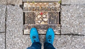 A person wearing blue shoes stands over a sewer grate.