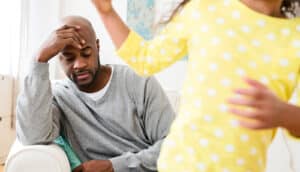 A father sits on a couch with his hand on his forehead while his daughter runs in the foreground.